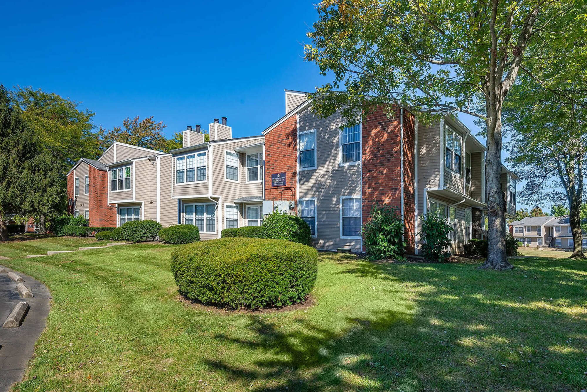 Exterior view of Steeplechase Apartments showing tan and brick-faced two-story buildings with manicured lawns, shrubs, and mature trees under a clear blue sky.