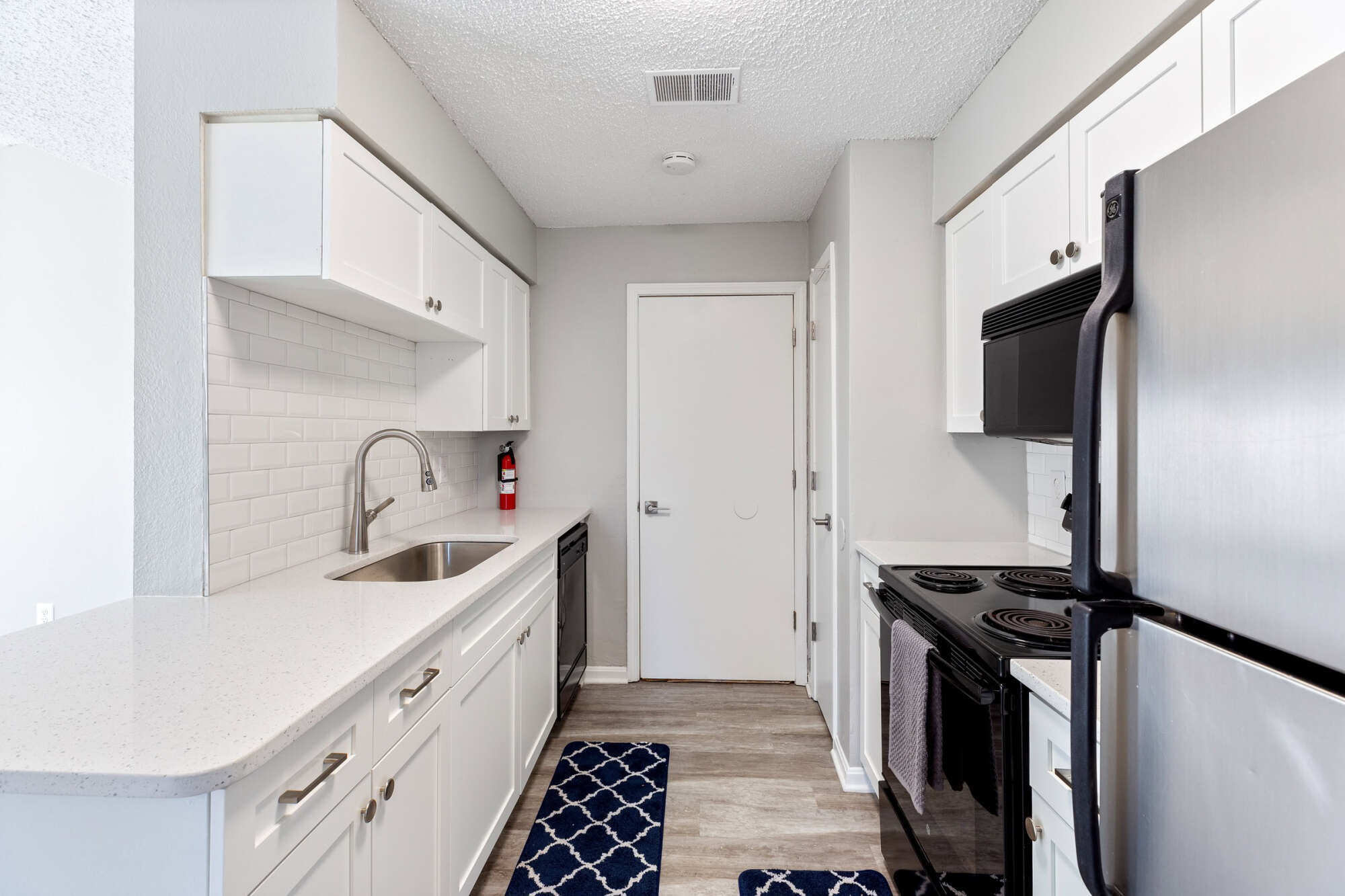 Bright galley kitchen with white shaker cabinets, light quartz countertops, subway tile backsplash, stainless steel refrigerator, and black appliances on wood-look flooring.