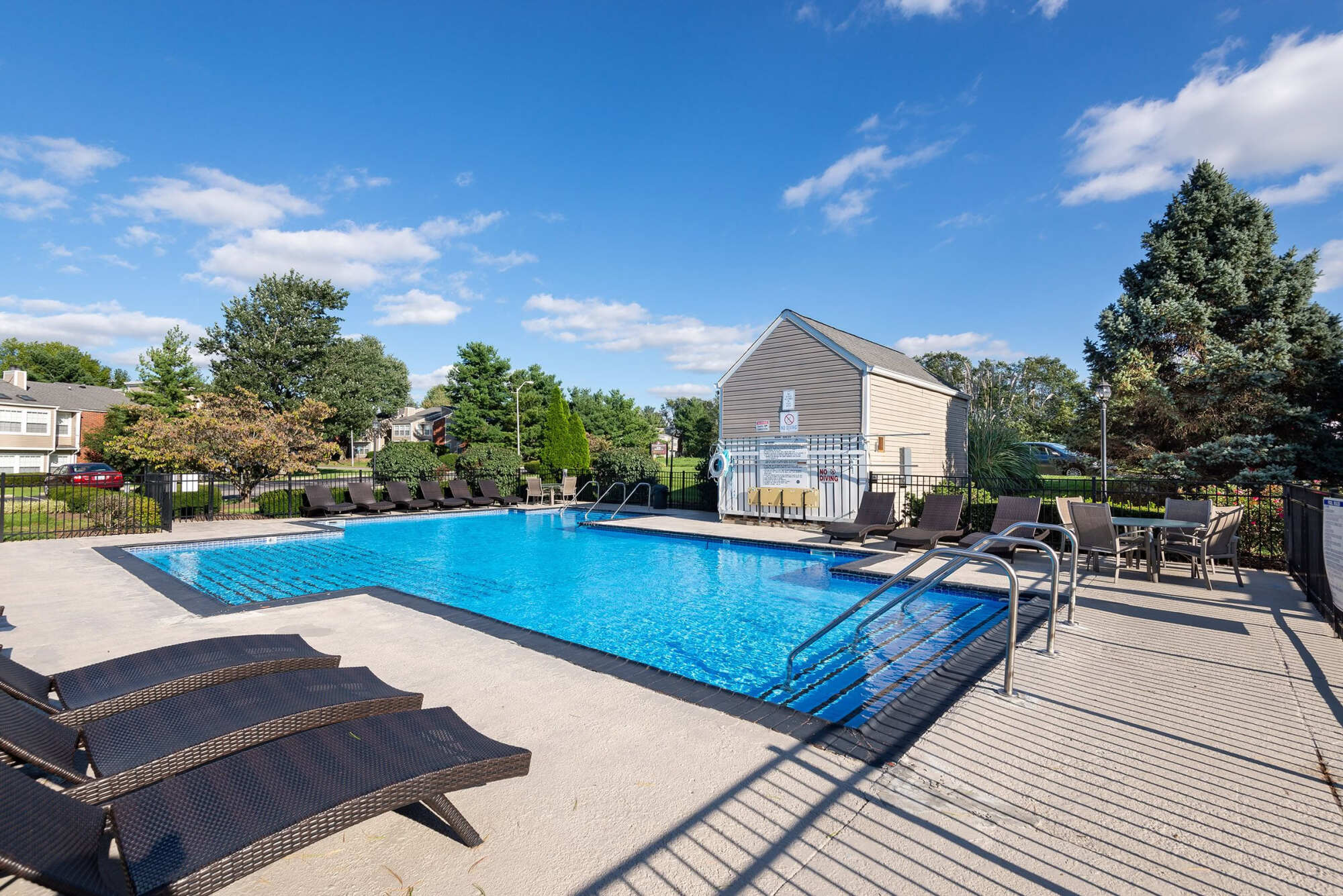 Outdoor swimming pool with blue water, surrounded by a sun deck, lounge chairs, a pool house, and landscaping under a partly cloudy sky.