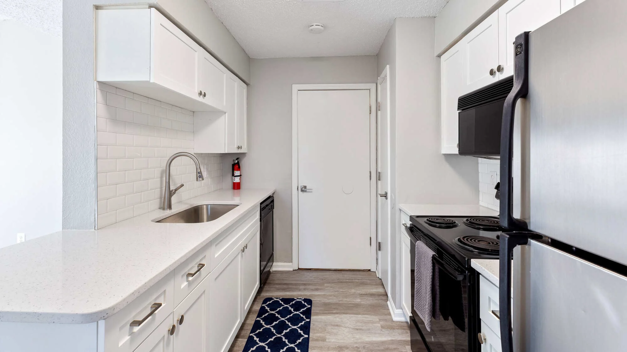 Renovated galley kitchen featuring white cabinetry, quartz countertops, white subway tile backsplash, stainless steel refrigerator, and black range and dishwasher.